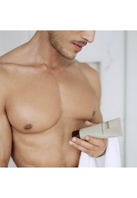Gel detergente per il viso da uomo in una confezione beige con cappuccio marrone scuro, tenuto da una persona con pelle chiara e un asciugamano appoggiato sul braccio.