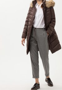 Bruine pufferjas met een met bont gevoerde capuchon over een witte geknoopte shirt, gecombineerd met grijze op maat gemaakte broek en zwarte gepatineerde leren schoenen.