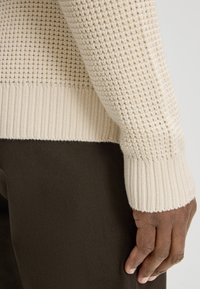 Dzianinowy sweter w kolorze kremowym z teksturowanym wzorem, z prążkowanymi mankietami i dołem, noszony na ciemnobrązowym materiale. Zbliżenie na rękaw i dłoń.