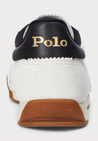 Vit mockasinsneaker med häl i svart läder, guld "Polo"-logotyp och brun gummisul. Minimalistisk design med släta texturer.