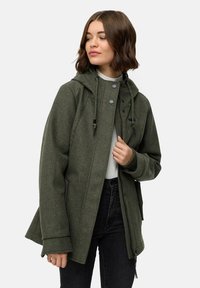 Femme portant une veste à capuche olive avec cordons, un haut blanc et un jean noir, regardant légèrement vers sa gauche.