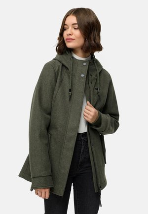 Vrouw in een olijfgroene hoodie met koorden, witte top en zwarte spijkerbroek, die iets naar links kijkt.