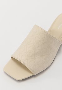 Sandale à glissière beige avec une tige en tissu texturé présentant un motif en relief. Design à bout ouvert et semelle plate lisse pour le confort.