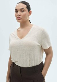 T-shirt beige à col en V en tissu léger, légèrement transparent. Coupe ample avec des manches courtes, associé à un pantalon taille haute marron foncé.