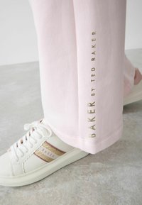 Rosa sweatpants med en slät textur och guldtext som läser "BAKER BY TED BAKER." Parat med vita sneakers med guld- och rosa detaljer.