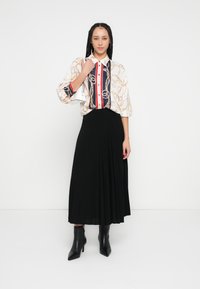 Bluza s ovratnikom s uzorkom lanaca i pruga, uparen s crnom plisiranom suknjom. Model nosi crne gležnjače i drži malu bijelu torbicu.