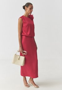 Red sleeveless dress with a flower accent, mid-calf length. Additional details include a cream handbag and white heeled sandals.