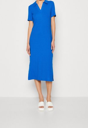 Kobieta ubrana w niebieską prążkowaną sukienkę za kolano z krótkim rękawem i kołnierzykiem, nosząca białe sandały na platformie, stojąca na jednolitym tle.