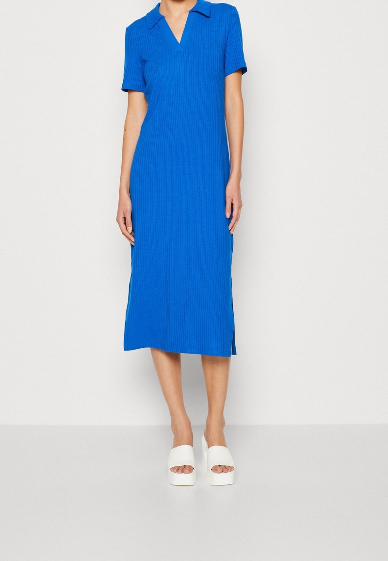 Femme portant une robe coloris bleu vif, côtelée, à manches courtes et longueur genou, avec un col, et des sandales blanches à plateforme, debout devant un fond uni.