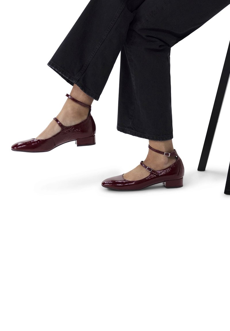 Scarpe in pelle verniciata color bordeaux con tacco basso a blocco, cinturini alla caviglia e design della punta arrotondata, indossate ai piedi di una persona seduta.