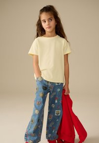 Mladá dívka s dlouhými vlasy, oblečená v bledě žlutém tričku a barevných džínách s vzorem, držící červenou mikinu na jednobarevném pozadí.