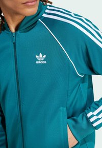 Tyrkysová zipová bunda s bílým logem adidas. Obsahuje bílé pruhy na ramenou a rukávech. Hladká, atletická tkanina, kapsy po stranách.