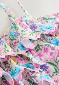 Virágmintás fürdőruha, rózsaszín, kék és sárga virágokkal fehér alapon. Ráncolt szegélyekkel és vékony, állítható pántokkal rendelkezik.