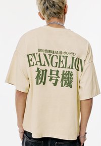 Beige, oversized póló zöld "EVANGELION" szöveggel és japán karakterekkel a hátsó részén, puha pamut anyagból készült. Rövid ujjú.