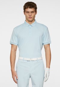 Ljusblå polo skjorta i ett mjukt tyg med korta ärmar, med en knappslå och logodetaljer på kragen och ärmen. Vit bälte och handskar.