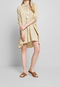 Robe jaune rayée avec un design en étages, un col rond et des accents en franges. Associée à des sandales plates marron et des manches ajustées.