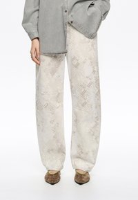 Pantalons évasés clairs à motifs avec une surface texturée, associés à des chaussures en daim marron et une chemise en denim grise.