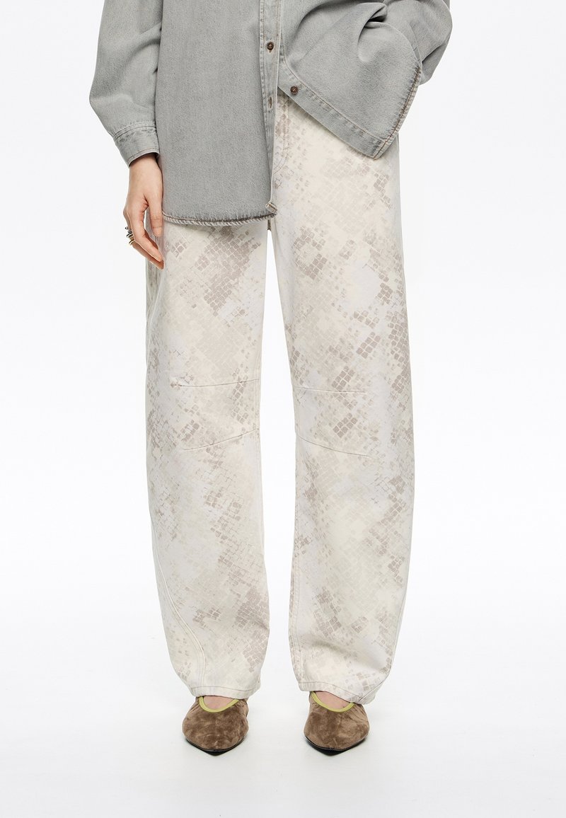 Pantalons évasés clairs à motifs avec une surface texturée, associés à des chaussures en daim marron et une chemise en denim grise.