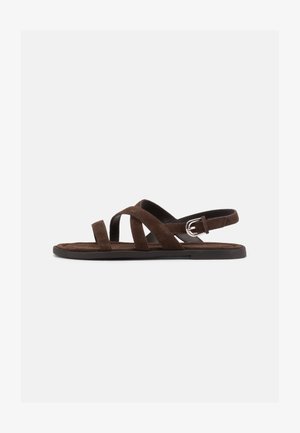 Hnedá semišová plochá sandála s tromi úzkymi pásikmi prekrývajúcimi nohu a strieborným prackovým remienkom na členku na bielom pozadí.