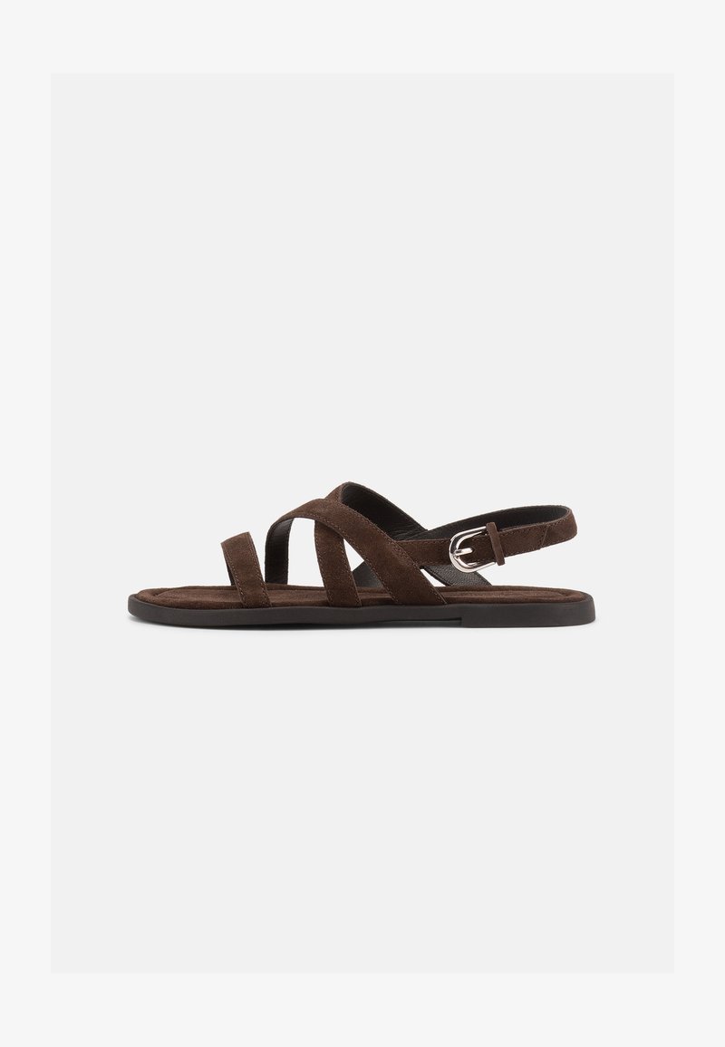 Καφέ σουέντ φλατ σανδάλι με τρεις λεπτές λωρίδες που διασχίζουν το πόδι και ασημένια αγκράφα στον αστράγαλο, σε λευκό φόντο.