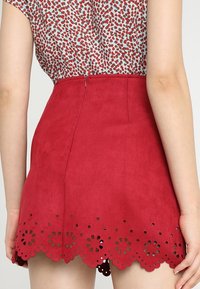 Mini-jupe rouge en suédine synthétique avec un ourlet festonné présentant des découpes florales. Fermeture éclair au dos et texture lisse.