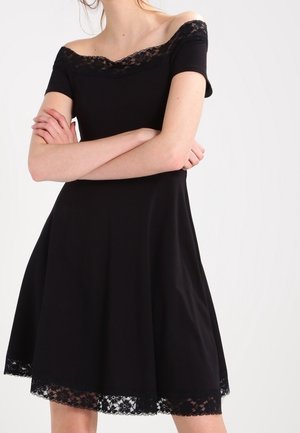 Žena v krátkých černých šatech bez ramínek s krajkovou ozdobou na výstřihu a lemu, s rukama zkříženýma přes hrudník.