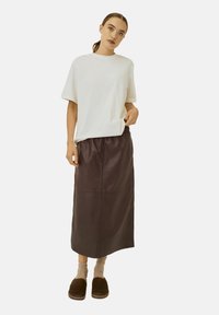 T-shirt blanc à manches courtes, jupe en cuir marron jusqu'aux chevilles, pantoufles marron, portant des chaussettes beiges. Design minimaliste et coupe décontractée.