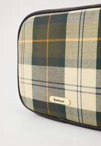 Saco de tecido xadrez em tons de verde, bege e laranja com uma placa em dourado do logo "Barbour". Apresenta uma textura suave e bordas arredondadas.