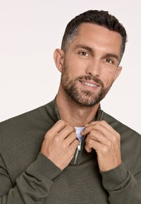 Donkergroene ritssluiting trui met geribbelde manchetten, halve ritssluiting en textuurstof. Het model houdt de ritssluiting met beide handen vast en toont een gelaagd uiterlijk.