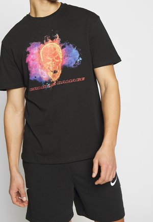 Mies, jolla on musta T-paita, jossa on värikäs liekehtivä pääkallo ja teksti "CRIMINAL DAMAGE", yhdistetty mustiin Nike-shortseihin.