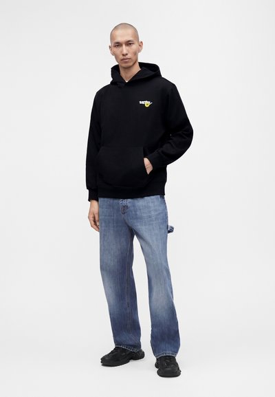 Homme portant un sweat à capuche noir avec un petit smiley jaune et un texte, jean bleu clair et baskets noires, debout avec une main dans la poche.