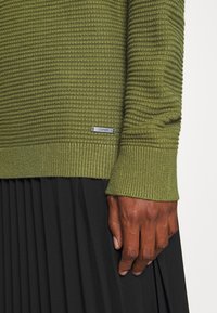 Maglione verde oliva strutturato con polsini a coste, dotato di una piccola etichetta logo argentata. Indossato con una gonna nera plissettata.