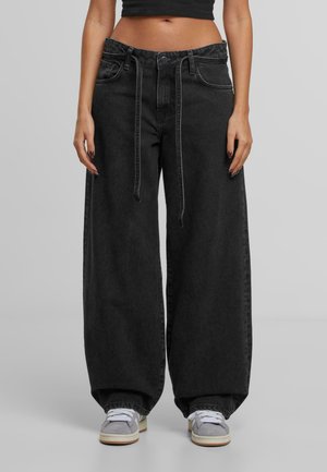 Moteris vilkinti juodas plačias kelnes su priekyje surišama dirželiu ir pilkais sportbačiais, stovinti prieš neutralią pilką foną.