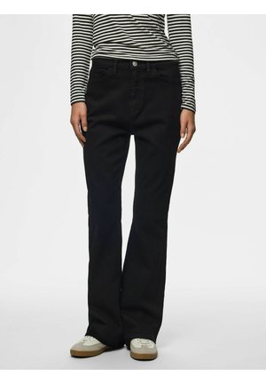 Personne portant un pantalon noir évasé, une chemise à manches longues rayée et des baskets beiges, se tenant devant un fond uni.