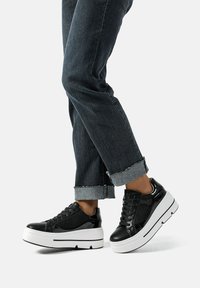 Sneakers platform neri con dettagli in rete e pelle verniciata, caratterizzati da una suola bianca e accenti testurizzati. Indossati con jeans scuri con risvolto.