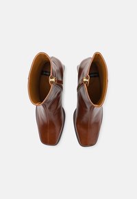 Botas de tornozelo em couro castanho com um design elegante, bico quadrado, detalhes em dourado e forro em tom de areia contrastante. Textura lisa e brilhante.