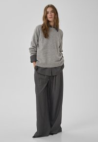 Grauer, gestrickter Pullover mit geripptem Saum, darüber getragen ein dunkles, gestreiftes Hemd mit Knöpfen und lockere, weit geschnittene, gestreifte Hosen.