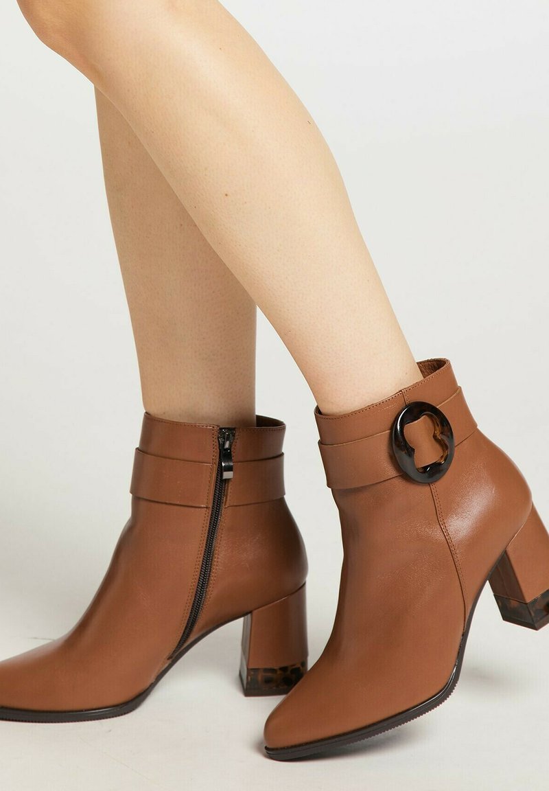 Braune Leder-Ankle-Boots mit spitzer Zehenform, Reißverschluss und einem runden schwarzen Akzent. Das klobige transparente Absatzdesign verleiht Textur.