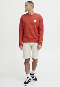 Rode sweatshirt met een ronde halslijn en een klein cirkelvormig logo op de borst, gecombineerd met beige shorts en zwarte sneakers.