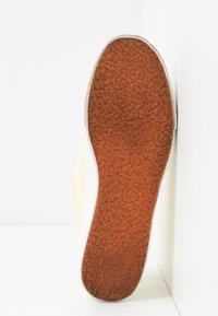 Baskets blanches reposant sur le côté montrant une semelle en caoutchouc marron texturée avec un motif ondulé et irrégulier et une semelle intermédiaire blanche.