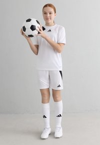 Balta sporta krekla un šortus ar melniem akcentiem, kombinēti ar baltām zeķēm un sporta apaviem. Tur rokās melnbaltu futbola bumbu.