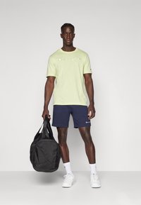 Camiseta de algodón verde claro con logo, pantalones cortos azul marino y una bolsa de deporte negra. El modelo lleva zapatillas blancas y calcetines con acentos de logo.