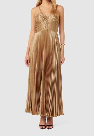 Kobieta ubrana w metaliczną złotą plisowaną sukienkę midi na cienkich ramiączkach, w parze z czarnymi sandałkami na otwartym palcu oraz złotą bransoletką na lewej ręce.