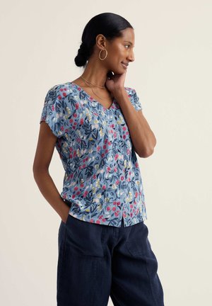 Sieviete ar tumšiem, sasietiem matiem valkā riņķveida auskarus, zilu ziedu raksta īsroku blūzi un tumšas bikses, pozējot ar vienu roku uz kakla.