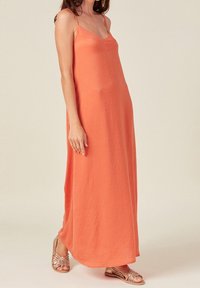Fließendes oranges Maxikleid mit dünnen Trägern und V-Ausschnitt. Es hat eine weiche Textur und eine lockere Silhouette und wird mit metallischen Sandalen kombiniert.