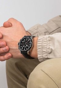 Orologio cronografo nero con accenti argentati e cinturino nero testurizzato, esposto su una mano chiusa sopra una giacca beige e pantaloni.
