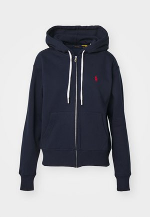 Laivastonsininen zip-up huppari, joka on valmistettu puuvillasekoitteesta. Vasemmassa rinnassa on punainen logo, valkoiset nyörit ja etutaskut. Sileä pinta.