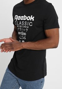 Musta puuvillapaita, jossa on valkoinen "Reebok CLASSIC" -grafiikka ja monikielistä tekstiä alapuolella. Pyöreä pääntie, lyhyet hihat.