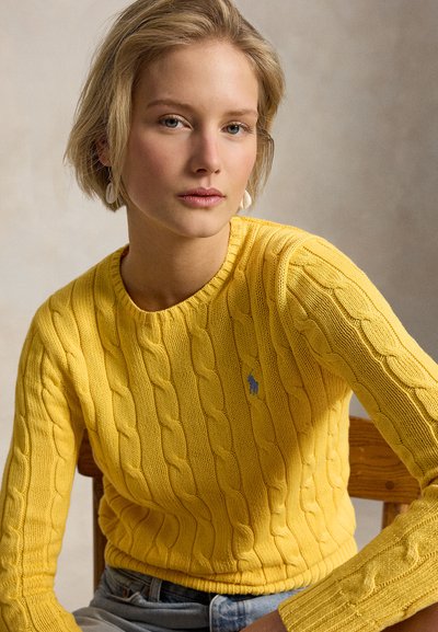 Νεαρή γυναίκα με κοντά ξανθά μαλλιά, φοράει κίτρινο πουλόβερ με κοτσίδες και ανοιχτόχρωμο τζιν, καθισμένη σε ξύλινη καρέκλα μπροστά σε ουδέτερο φόντο.