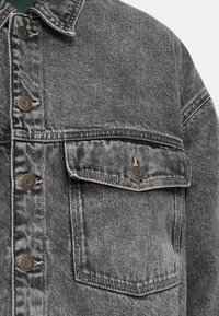 Close-up da parte da frente de uma jaqueta de denim cinza com botões de metal e um bolso de aba no peito.